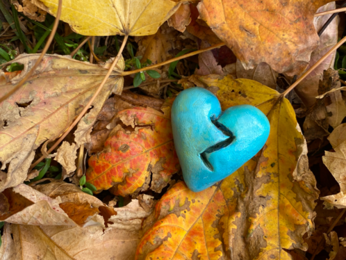 a small blue painted broken heart made of clay lays in a bed of fallen gold rotting leaves
