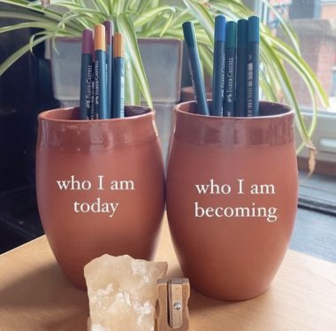 two terra cotta cups holding pencil crayons on a desk in front of a spider plant. One cup says "who I am today" and the other says "who I am becoming"